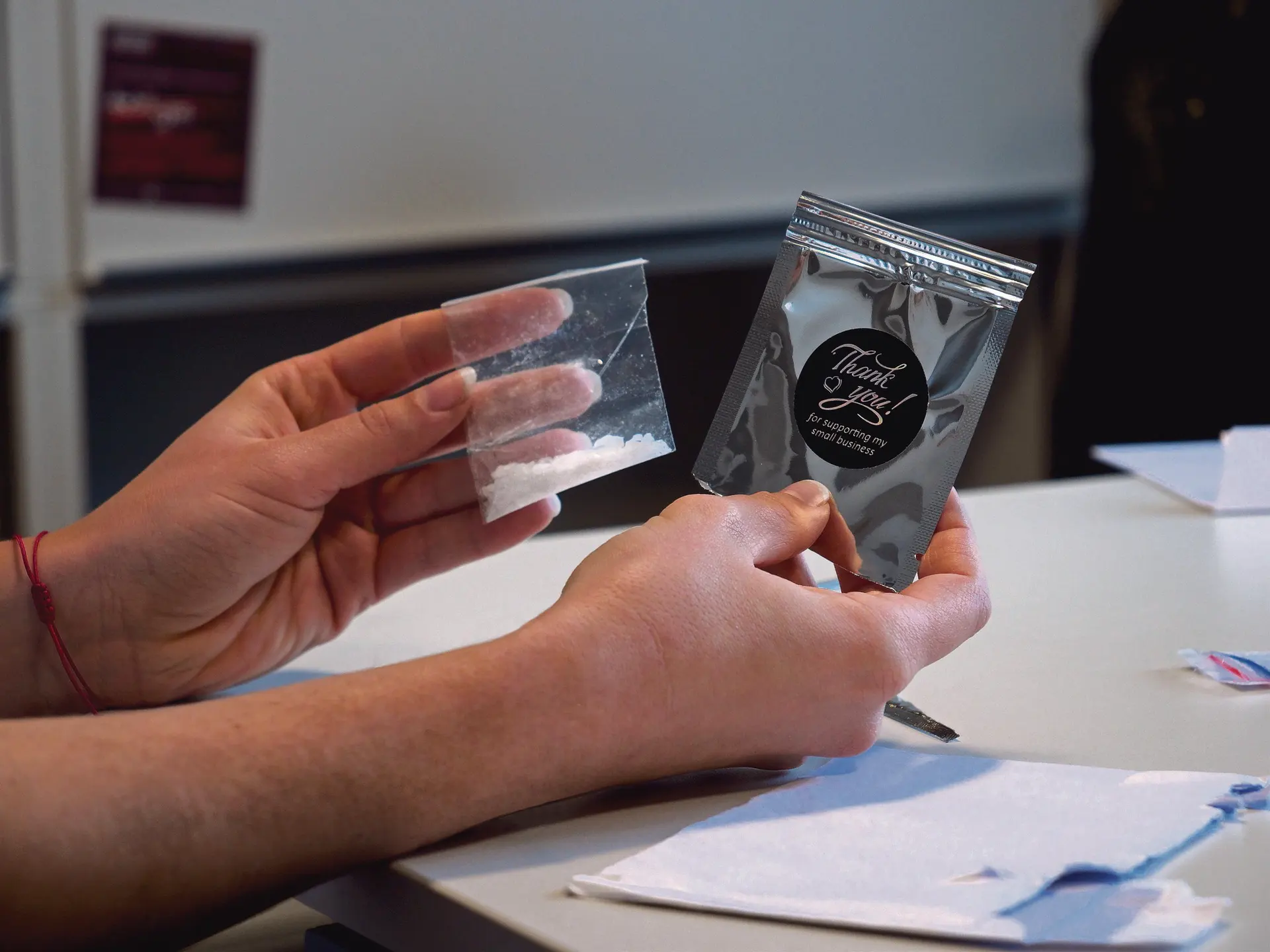 Two hands hold a small plastic bag of white powder and a foil pouch with a 'Thank you' sticker.
